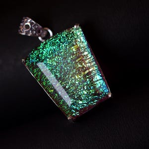 May include: A rectangular pendant featuring a vibrant, iridescent green and gold textured surface with a metallic sheen. The piece is encased in a silver-toned metal setting with a decorative, textured bail at the top for attachment to a chain.