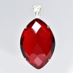 May include: A marquise-cut deep red gemstone pendant set in a silver-toned metal prong setting. The faceted stone features a pointed top and bottom with a reflective surface, suspended from a decorative silver-toned metal bail at the top.