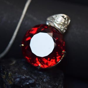 May include: A round, faceted deep red gemstone pendant set in a silver-toned metal prong setting with an ornate, filigree-style bail. The pendant is attached to a thin, silver-toned chain, showcasing a brilliant, reflective surface.