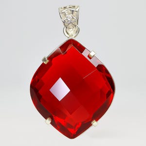 May include: A large, cushion-cut, vibrant red gemstone pendant set in a silver-toned metal prong setting. The faceted stone features a brilliant, transparent finish with a reflective surface. A decorative, ornate silver bail sits at the top.