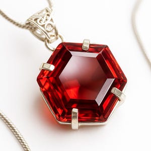May include: A hexagonal, deep red gemstone pendant set in a silver-toned metal prong setting. The pendant is attached to a silver-toned chain with a decorative, filigree-style bail at the top, showcasing a faceted surface and clear red color.