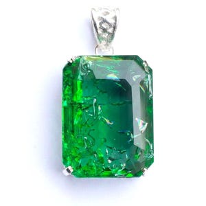Pode incluir: Um pendente retangular verde-esmeralda com superfície facetada, texturas internas rachadas e inclusões iridescentes. A gema é fixada por quatro garras de prata e ligada a uma argola de filigrana de prata decorativa no topo.