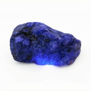 May include: A natural, unpolished sapphire. The sapphire is a rich, dark blue with a rugged, textured appearance. The gemstone is set against a white backdrop, emphasizing its natural colour and shape.