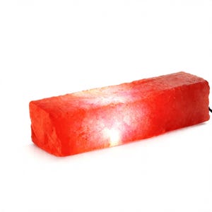 May include: A rectangular Himalayan salt lamp with a rough, crystalline texture and a vibrant orange-red glow. The block-shaped lamp emits a warm light from within, highlighting the natural mineral variations and jagged edges of the salt surface.