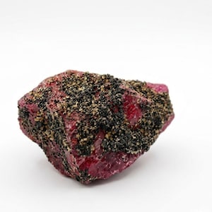 May include: A raw, unpolished mineral specimen featuring a deep magenta-red crystalline base. The surface is partially covered with a dense layer of small, dark black and metallic bronze-colored granular inclusions, creating a textured contrast.