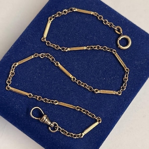 Puede incluir: Una cadena de reloj de bolsillo dorada con un cierre y un anillo, sobre una superficie azul. La cadena presenta segmentos alternos de pequeños eslabones y barras rectangulares más largas. El cierre y el anillo son plateados.