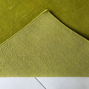 K&ouml;nnte beinhalten: Ein lindgr&uuml;ner Teppich mit einer umgeklappten Ecke, die die Unterseite zeigt. Die Oberseite hat eine dichte Kurzflor-Textur, w&auml;hrend die R&uuml;ckseite ein strukturiertes, gitterartiges Webmuster im gleichen leuchtenden Gr&uuml;nton aufweist.