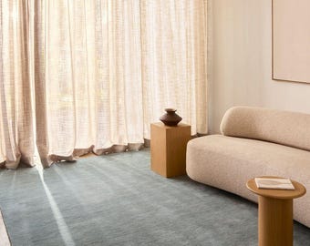 Könnte beinhalten: Ein Innenraum mit einem großen, einfarbigen schieferblauen Teppich, der den Boden bedeckt. Ein beiges, geschwungenes Sofa steht auf dem Teppich, ergänzt durch einen zylindrischen Holztisch, einen quadratischen Holzsockel mit einer braunen Vase und Vorhänge.