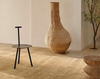Könnte beinhalten: Ein schwarzer Metallstuhl mit runder Sitzfläche und T-förmiger Rückenlehne, eine hohe, geflochtene, hellbraune Bodenvase mit bauchiger Basis und engem Hals sowie ein zylindrischer Holzhocker mit geschnitzter Textur auf einem beigen Teppich.