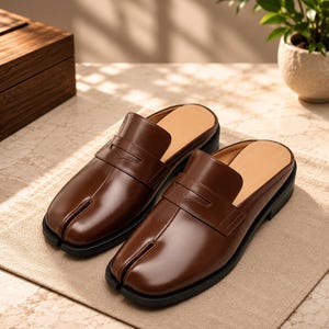 May include: A pair of brown leather backless loafers featuring a distinctive split-toe design and a classic penny loafer strap across the vamp. These slip-on shoes have a smooth, polished finish, a tan interior lining, and a black rubber sole.