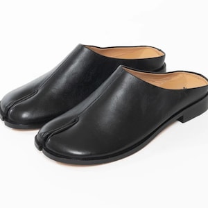 Puede incluir: Un par de mules sin talón de cuero negro con un diseño distintivo de punta dividida. Estos zapatos tienen una parte superior de cuero negro liso, un tacón bajo y un forro interior de color tostado. Una costura recorre el centro del empeine.