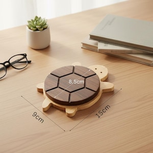 May include: A wooden turtle-shaped coaster with a dark brown segmented top and a light brown base. The coaster measures 15 cm long and 9 cm wide, with the segmented top measuring 8.5 cm across. A succulent plant and books are in the background.