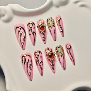 May include: Pink artificial nails with gold flame designs and heart-shaped gems. The nails are almond-shaped and arranged on a white decorative plate. Some nails have clear gems.