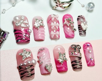 May include: A collection of ten artificial nails in various designs. The nails feature pink and white gradients, zebra stripes, bows, and star-shaped embellishments. Some nails have pearl and rhinestone accents. A silver starfish ring and beaded jewelry are also present.
