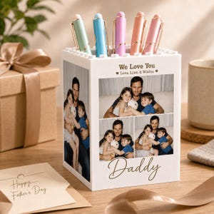 May include: A white square pen holder featuring a top edge with a building block texture. The sides are printed with three family photographs and the text "We Love You Love, Liam & Wislim" and "Daddy". Five pastel-colored pens are inserted inside.