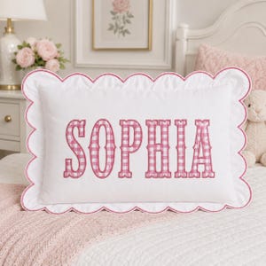 May include: A rectangular white pillow featuring a scalloped edge trimmed in pink. The center of the pillow displays the name SOPHIA embroidered in a pink and white gingham pattern. The fabric is smooth and the design is centered on the pillow.
