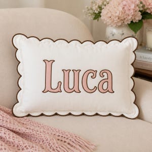 May include: A rectangular white throw pillow featuring a scalloped edge trimmed in dark brown. The center of the pillow displays the name Luca embroidered in a soft pink font with a dark brown outline, centered on the white fabric surface.