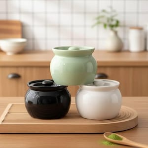 Può includere: Tre barattoli di ceramica con coperchi in un ambiente cucina. I barattoli sono verde chiaro, neri e bianchi, e poggiano su un vassoio di legno. Un cucchiaio di legno con polvere verde è in primo piano.