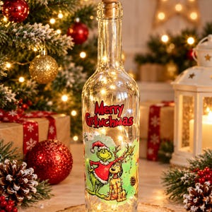 May include: A clear glass bottle with a cork stopper, illuminated by internal string lights. The bottle features a graphic of a green character in a red suit standing next to a dog with antlers, with the text Merry Grinchmas printed above.
