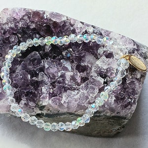 May include: A clear crystal beaded bracelet with a gold charm that says "Forever Yours". The bracelet is displayed on a purple amethyst geode.