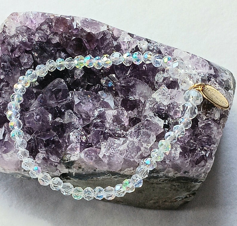 May include: A clear crystal beaded bracelet with a gold charm that says "Forever Yours". The bracelet is displayed on a purple amethyst geode.