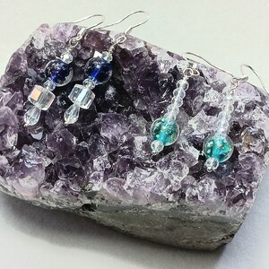 May include: Three pairs of dangle earrings with silver hooks and crystal beads. The earrings feature blue and teal glass beads with iridescent accents. The earrings are displayed on a purple amethyst geode.