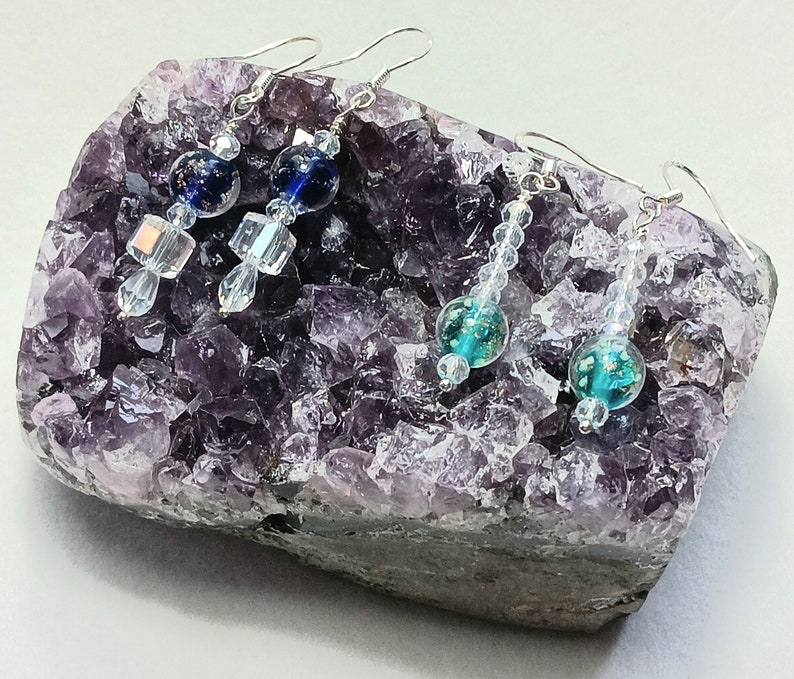 May include: Three pairs of dangle earrings with silver hooks and crystal beads. The earrings feature blue and teal glass beads with iridescent accents. The earrings are displayed on a purple amethyst geode.
