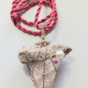 May include: A pink crystal pendant with gold wire wrapping, a pearl, and a pink gemstone. The pendant is attached to a red cord necklace.