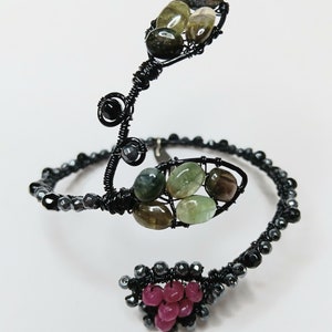 May include: A black wire wrapped bracelet featuring green and brown stones, and a cluster of pink stones. The bracelet is designed in a spiral shape.