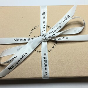 May include: A brown cardboard box with a white ribbon tied in a bow. The ribbon has the text "Navenadia" printed on it.