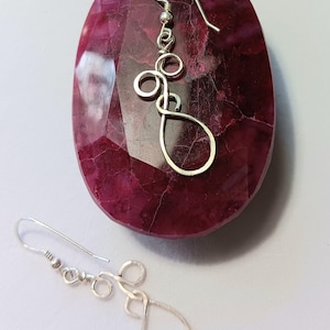 May include: A pair of silver dangle earrings with a teardrop shape and multiple small circles. The earrings are displayed on a red stone.