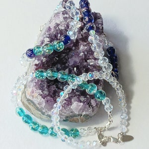 May include: Three strands of beaded necklaces, one clear, one turquoise with gold flecks, and one dark blue with gold flecks, displayed on a purple amethyst geode.
