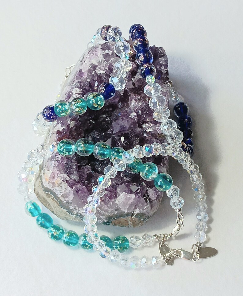 May include: Three strands of beaded necklaces, one clear, one turquoise with gold flecks, and one dark blue with gold flecks, displayed on a purple amethyst geode.