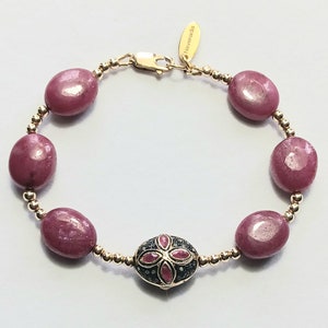 May include: A gold bracelet featuring a pink oval-shaped bead and a central piece with a flower design crafted from red rubies and black diamonds. The bracelet is made by the brand "Navananda".