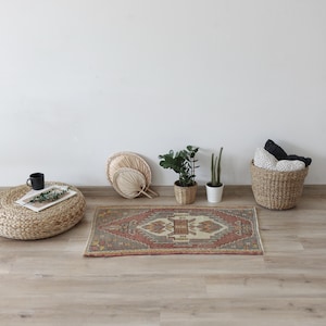 May include: A small, rectangular rug with a central geometric design in shades of red, beige, and gray. The rug is placed on a light-colored wooden floor, surrounded by decorative items like plants, fans, and a woven basket, creating a cozy, bohemian aesthetic.