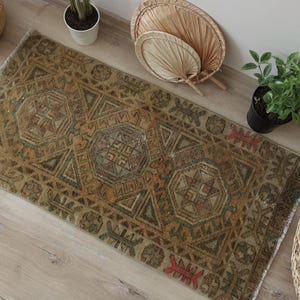 Puede incluir: Una alfombra rectangular con una paleta de colores desvaídos y apagados en verde oliva, marrón y ocre. La alfombra muestra un patrón geométrico repetitivo con tres medallones octogonales centrales, bordes intrincados y pequeños detalles en rojo.