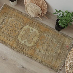 Puede incluir: Una alfombra rectangular que presenta una paleta de colores amarillo mostaza descolorido y beige con intrincados patrones geométricos y un diseño de medallón central. La alfombra tiene una textura de pelo corto y un aspecto desgastado.