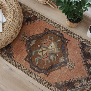 May include: A rectangular, vintage-style rug with a central medallion design in shades of brown, blue, and beige. The rug has a distressed appearance and a border with geometric patterns. The rug is placed on a wooden floor, with a woven ottoman and plants nearby.