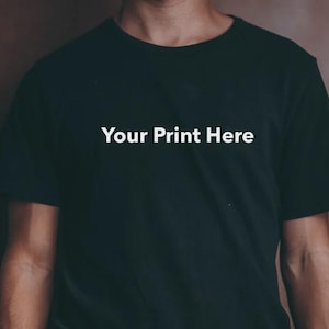 Puede incluir: Camiseta negra con el texto blanco "Your Print Here". La camiseta es de cuello redondo y parece estar hecha de un material suave. El fondo es de color marrón liso.