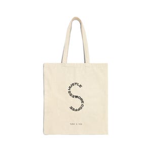 May include: Cream-colored tote bag with black text. The text forms a stylized "S" shape with the words "Hustle More Coffee" and "take a sip" at the bottom. The bag has two long handles.