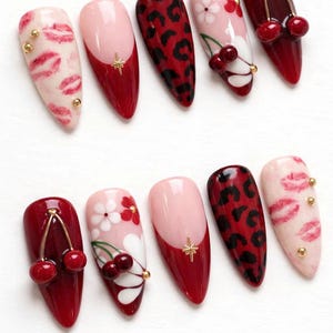 May include: A set of ten almond-shaped artificial nails with various designs. The designs include red leopard print, pink and red cherry accents, pink and white floral patterns, and pink with red tips. Some nails feature gold star or stud embellishments.