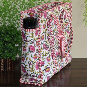 Puede incluir: Un bolso tote blanco con un estampado floral en rosa, amarillo y verde. El bolso tiene ribetes y asas rojas, y un bolsillo lateral que contiene una botella negra. El texto "BohaBlocks" es visible.