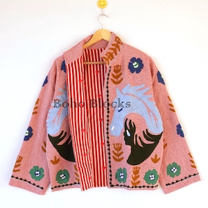 Può includere: Giacca rosa con design a cavallo e dettagli floreali. La giacca ha un interno a righe rosse e bianche e il testo "Boho Blocks". La giacca è appesa a una gruccia di legno.