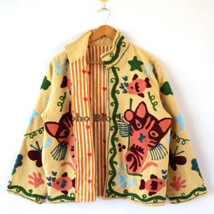 Può includere: Giacca beige ricamata con disegni colorati di pesci, gatti e fiori. La giacca ha un interno a righe arancioni e bianche e il testo "boho Block". La giacca è appesa a una gruccia di legno.