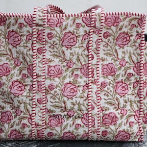 Puede incluir: Un bolso tote grande con un estampado floral. El bolso presenta un fondo blanco con flores rosas y hojas verdes. Los bordes y las asas están ribeteados con una tela a rayas burdeos y blancas. El bolso tiene un bolsillo lateral y el texto "Boho blocks" es visible.