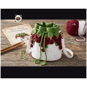 May include: A white crochet drawstring bag with a green top and red rose accents. The bag has a white strap and gold-colored hardware. A book titled "Rose Petal Drawstring Bag" and yarn are in the background.