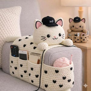 May include: A cream-colored, crochet cat-shaped sofa arm organizer with black heart accents. The cat wears a black cap with "H-P" embroidered on it. The organizer has multiple pockets for remote controls, phones, and notepads. A ball of pink yarn and a crochet hook are in one pocket.