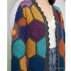 Peut inclure: Un cardigan crocheté à la main, aux couleurs vives, avec un motif géométrique unique. Le pull présente une variété de formes hexagonales dans des tons de violet, orange, bleu et sarcelle, avec une bordure festonnée grise. Le texte "Loomed & Hooked" est visible.