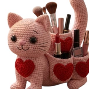 May include: A handmade, light pink crochet cat-shaped organizer with red heart accents. The cat has large black eyes and holds makeup brushes, lipsticks, and other beauty products in its pockets. A unique and charming storage solution.
