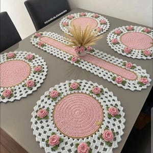 May include: A crocheted table setting featuring a pink and white color scheme. The set includes placemats and a table runner, all adorned with pink rose embellishments. The placemats have a diameter of approximately 12 inches.